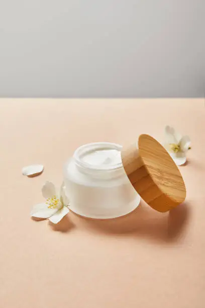 Photo of open jar with cream and wooden cap and few jasmine flowers on beige isolated on grey