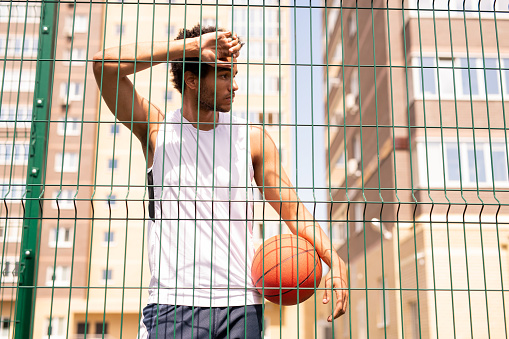 Adolescent basketball player in activewear leaning against fence of the court while having rest after training