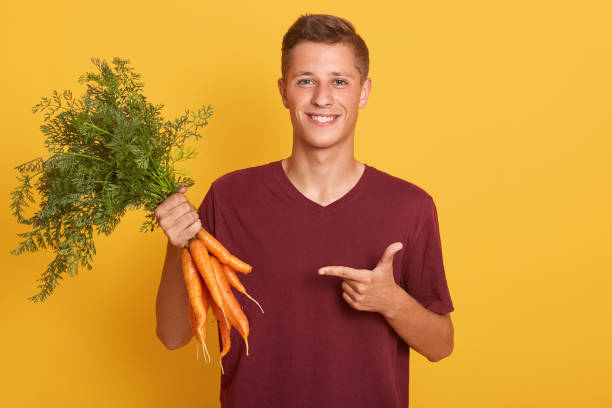 alimento cru e conceito saudável da alimentação. menino bem olhando de sorriso no grupo ocasional da terra arrendada da camisa de borgonha t das cenouras, apontando nela com o indicador sobre o fundo amarelo. espaço da cópia para o advertisment. - indexfinger - fotografias e filmes do acervo