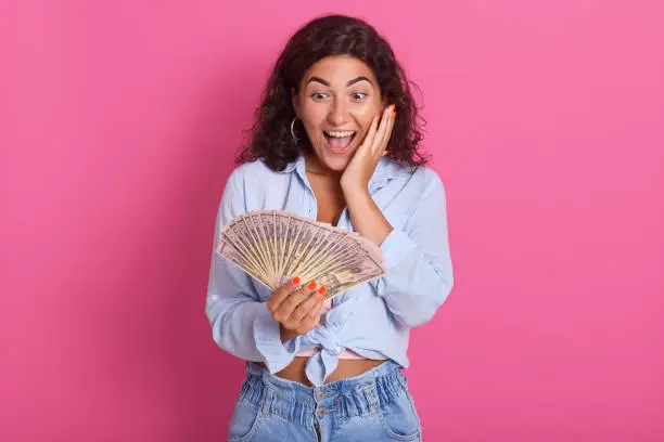 Image of beautiful brunette woman dressed in blue shirt and jeans, smiling and holding bunch of money bills isolated over pinkbackground, keeps mouth opened, has astonished facial expression.