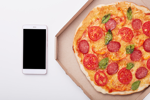 Pizza en caja y smartphone con pantalla en blanco sobre mesa blanca, vista superior de delicioso pepperoni aislado sobre fondo ligero. Teléfono móvil moderno cerca de la caja con pizza. Copiar espacio para publicidad. photo