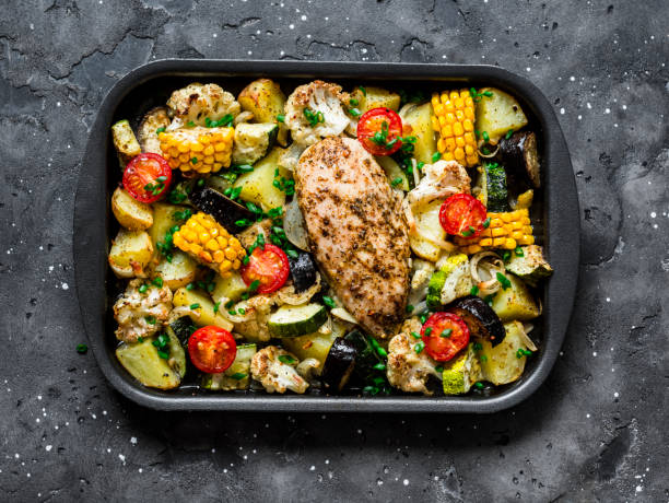 Chicken breast baked with potatoes, cauliflower, corn, eggplant, zucchini, cherry tomatoes on a baking sheet on a dark background, top view. Healthy food concept Chicken breast baked with potatoes, cauliflower, corn, eggplant, zucchini, cherry tomatoes on a baking sheet on a dark background, top view. Healthy food concept baking sheet stock pictures, royalty-free photos & images