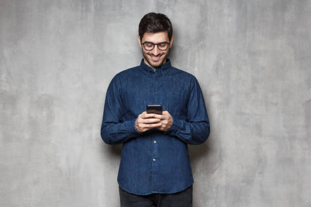 istock Internet surfing using smartphone. Portrait of young smiling man typing a message with new app on his phone, standing against gray textured wall 1164586372