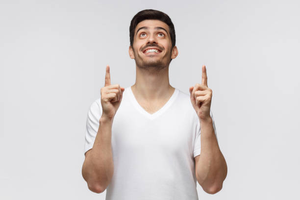 istock Handsome man in white t-shirt pointing up with both hands isolated on gray background 1164586486