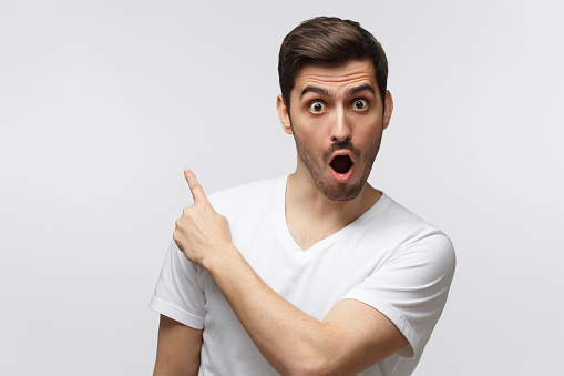 Young surprised man isolated on gray background in white t-shirt looking at camera with open mouth, pointing left, copyspace for ads