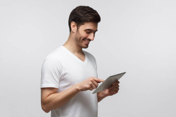 istock Young man looking at screen of tablet computer he is holding in hands in front of him, isolated on gray background 1164586492