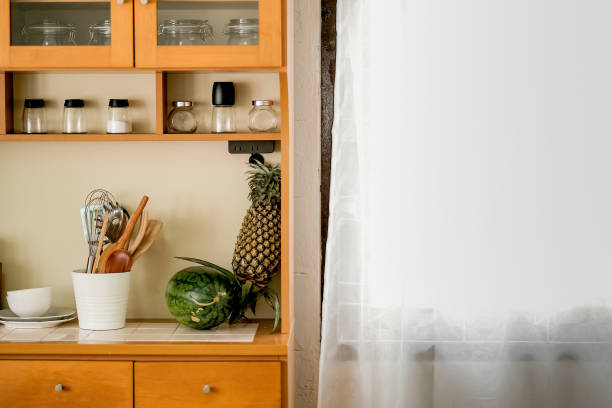 Part of kitchen area contain some accessories and fruit include water melon and pineapple in house near the window with white curtain during day time