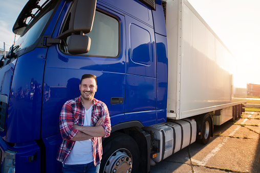 Portrait of middle aged Caucasian trucker in casual clothes standing in front of truck long vehicle with trailer. Transportation services.
