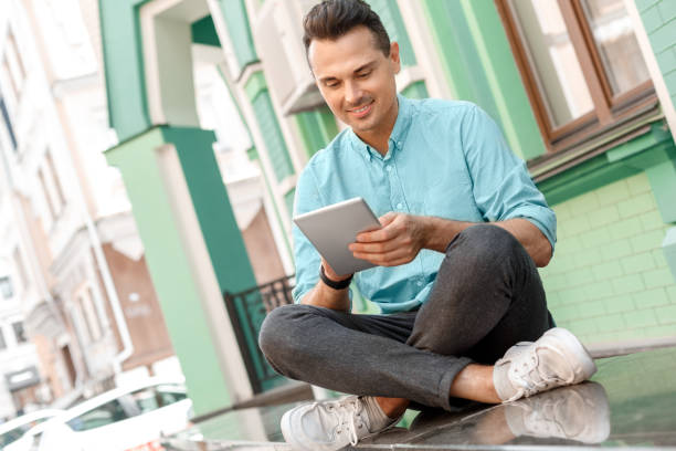 istock Outdoors leisure. Young man sitting on city street browsing digital tablet smiling joyful 1164612596