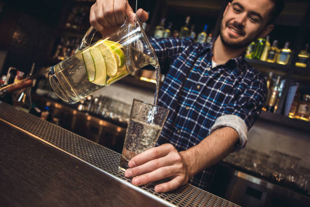 Young bartender standing at bar counter pouring lrmon cocktail into glass joyful