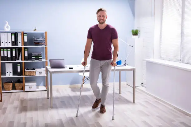 Handicapped Businessman Walking On Hardwood Floor With Crutches At Workplace