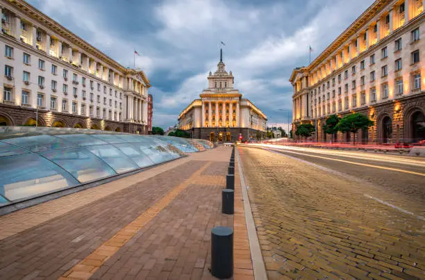 Panoramic long exposure photo from Sofia city centre in Bulgaria, Eastern Europe during twilight / rush hour with all the surrounding buildings night illumination. Including the iconic architecture: the old house of parliament, ministry and presidency next to the famous yellow pavement boulevard. Dragged shutter technique used to capture the blurred headlight/taillights from the road traffic. Shot on Canon full frame EOS R system for premium quality and resolution.