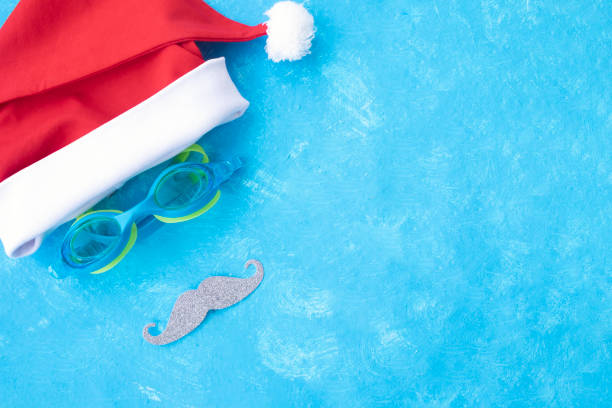Hat of Santa Claus with goggles for swimming and a mustache. Christmas vacation, sandals and swimming glasses by water, slippers and pool goggles near swimming pool stock photo