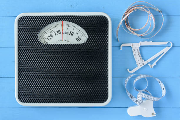 istock Mechanical weight scale, body mass control concept : Bathroom scale, personal accurate body fat tester / skin fold caliper measurement tool for stomach / belly and measuring tape on blue background 1164668669