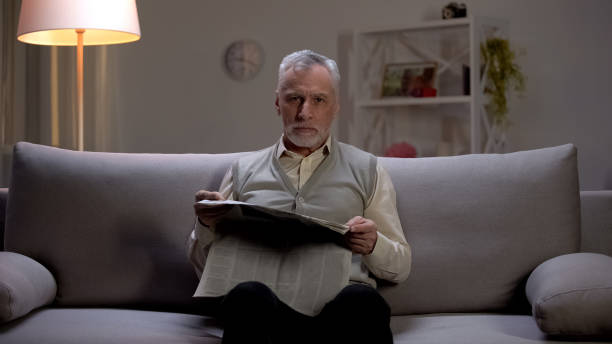 istock Old man reading newspaper in evening, seriously looking into camera, bad news 1164674230