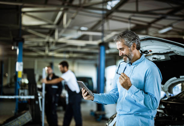 Happy mid adult manager working on touchpad in auto repair shop. Happy inspector using digital tablet in auto repair shop. There are people in the background. auto repair shop mechanic digital tablet customer stock pictures, royalty-free photos & images