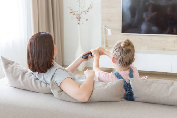 мать и дочь, сидя на диване смотреть телевизор, борьба за пульт дистанционного управления - family television fighting watching стоковые фото и изображения