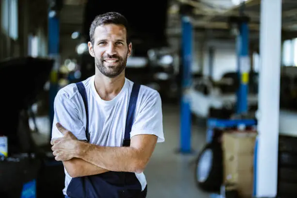 Young happy worker standing in auto repair shop with his arms crossed and looking at camera.