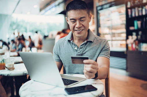 Smiling mid adult Asian ethnicity  man with laptop and credit card in cafe