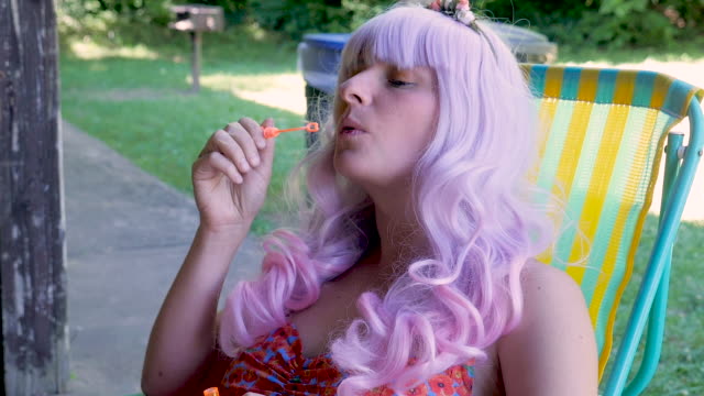 Young beautiful woman wearing a pink wig sitting in a lawn chair blowing bubbles