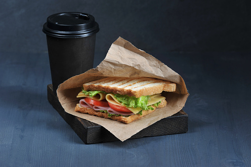 Sandwich with ham and cheese and paper cup with a drink on a dark background. Lettuce and tomato slices are used in the sandwich filling. The concept of fast food. Close-up. Macro shooting.