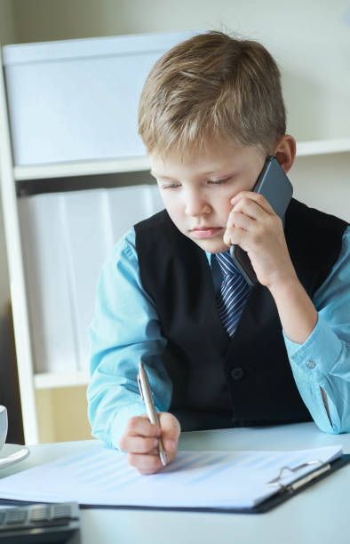 istock Little executive businessman boss boy making notes and talks on the phone and making notes sitting at the desk with laptop in office. Concept of early financial education 1164707931