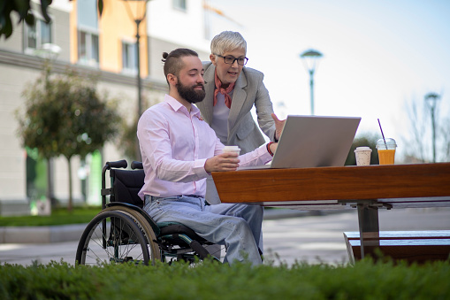 Young man in the wheelchair with the laptop having a business meeting with a mature gray haired woman in a public park.  The woman is standing behind him and looking at the laptop.