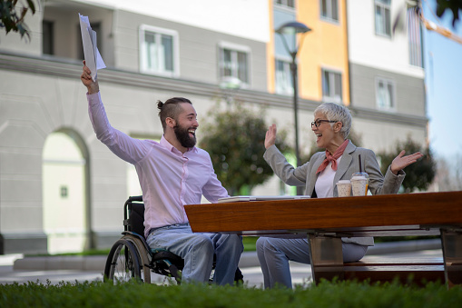 Mature grey haired woman and young man in the wheelchair with the laptop having a business meeting in a public park. They are celebrating.