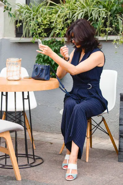 Brunette woman in sunglasses does makeup at a table in a cafe on a sunny summer day. Active style of the business woman and classical style in clothes.