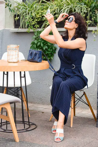 Brunette woman in sunglasses does makeup at a table in a cafe on a sunny summer day. Active style of the business woman and classical style in clothes.