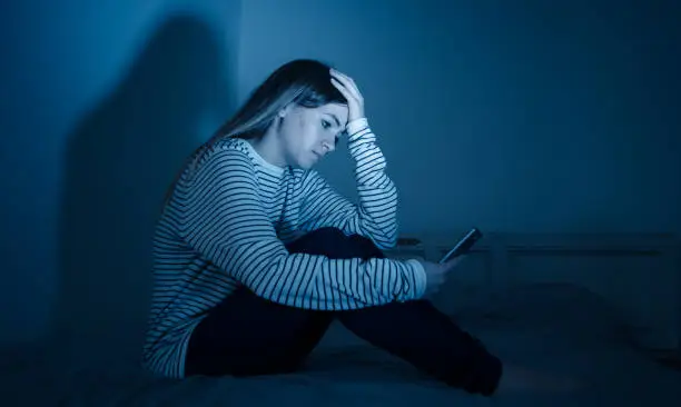 Sad desperate young teenager female girl on smart phone suffering from online bulling and harassment felling lonely and hopeless sitting on bed at night. Cyberbullying and dangers of internet concept.