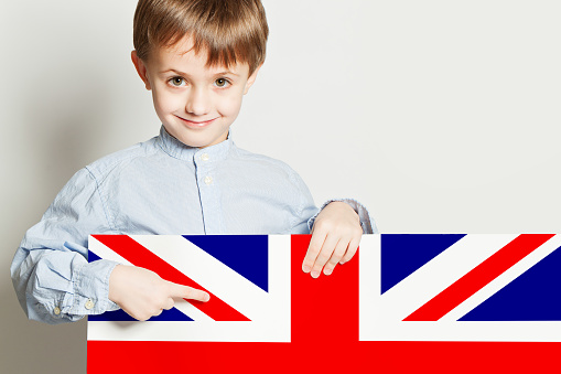 Little boy patriot standing side turned wrapped in a UK or British flag celebrates independence day expresses patriotism isolated on black background.