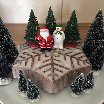Stock photo showing festive scene created by decorating a snowflake designed sponge cake covered in water icing with plastic figurines of Santa Claus, a snowman and Christmas trees.