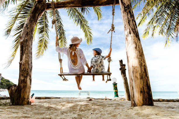 Rear view of carefree mother and son holding hands while swinging at the beach. Back view of mother and son enjoying in summer day while swinging at sandy beach in summer day. exotic vacations stock pictures, royalty-free photos & images
