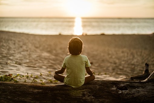 Back view of Zen-like kid practicing Yoga at the beach at sunset.