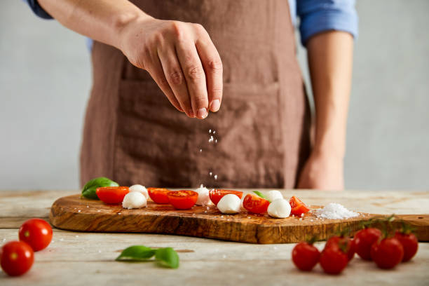 Adding pinch of salt Male cook is adding a pinch of salt on tomatoes, mozzarella and basil on a olive cutting board. salt seasoning stock pictures, royalty-free photos & images