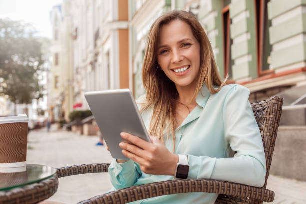 istock Young woman sitting at table in cafe drinking coffee browsing digital tablet looking camera cheerful 1164802520