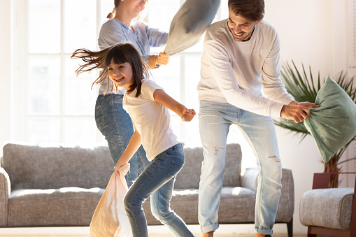 Playful young parents have fun with little preschooler daughter engaged in pillow fight in living room, happy family relax playing with small girl child, enjoy spending time at home together