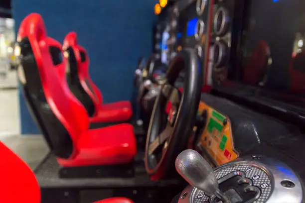 This closeup picture shows the gear shifter and steering wheel for a car racing video game at an arcade.  In the background of the picture are the seats used by video game players.