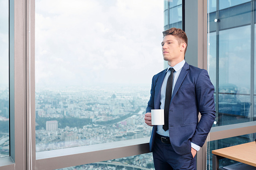 Serious businessman standing at big window in his corner office, drinking coffee and pondering over ideas for business development