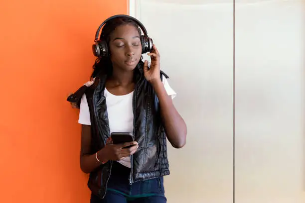 African Australian Teenage Girl Listening to Music Under Headphones While leaning Against Orange Wall Next To Elevator