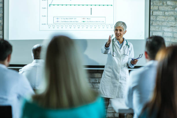 Happy senior doctor leading a presentation in a hospital. Happy mature female doctor talking to large group of her colleagues on a presentation in a hospital. medical education stock pictures, royalty-free photos & images