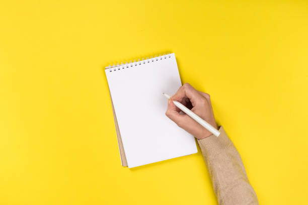 istock Woman's hands with perfect manicure holding pencil and  notepad as mockup for your design. Yellow background, flat lay 1164851963