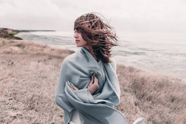 Photo of Attractive young woman standing on a windy cold beach wrapped in warm blanket with flower in her hand.