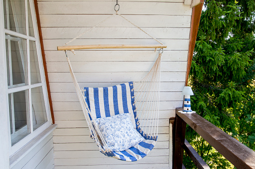 Blue and white striped pattern string and cotton hammock hanging chair, white painted wooden board background. Relaxing in countryside home garden balcony outdoors on summer day concept.