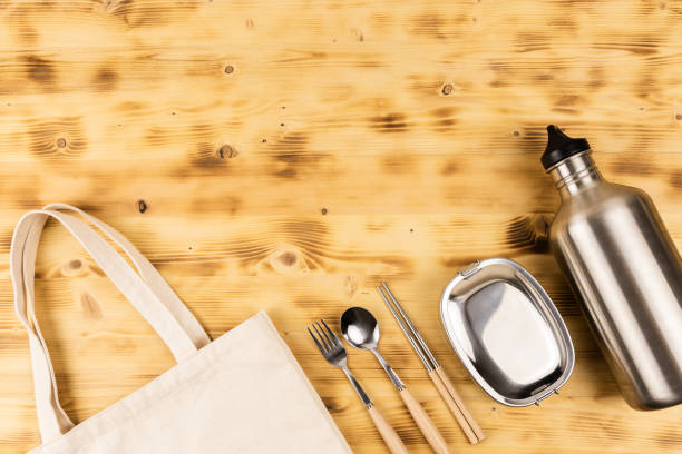 Stainless steel tableware; spoon, fork, chopsticks, lunch box and drinking water bottle lay on wooden table with eco friendly tote bag cut off. Flat lay with copy space.
