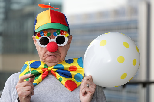 Sad faced man with Happy Birthday glasses, holding a small cake with a candle in it. Out of focus office buildings in the background.