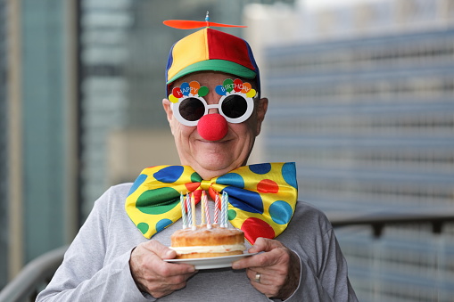 Smiling man in clown gear and Happy Birthday glasses holding a cake with candles. (It was yum!!)\nOut of focus office buildings in the background.