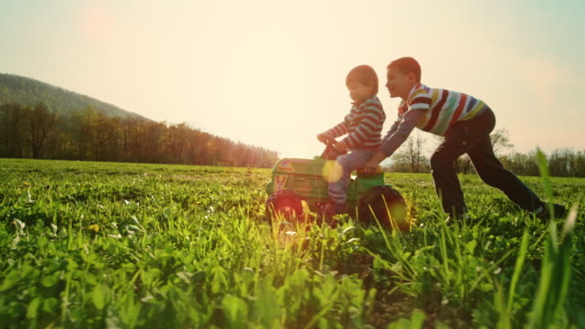SLO MO TS Boy pushing his younger brother on a toy tractor in a sunny meadow