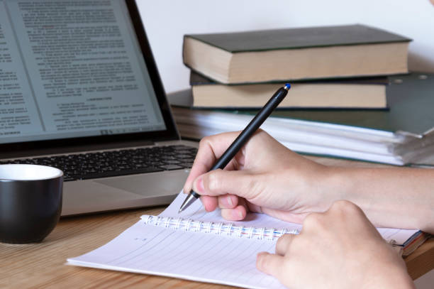 istock women's hands write with a pen in a notebook, on the table is a laptop, books and a cup. Working process. Study with books and laptop. Note-taking 1164910356